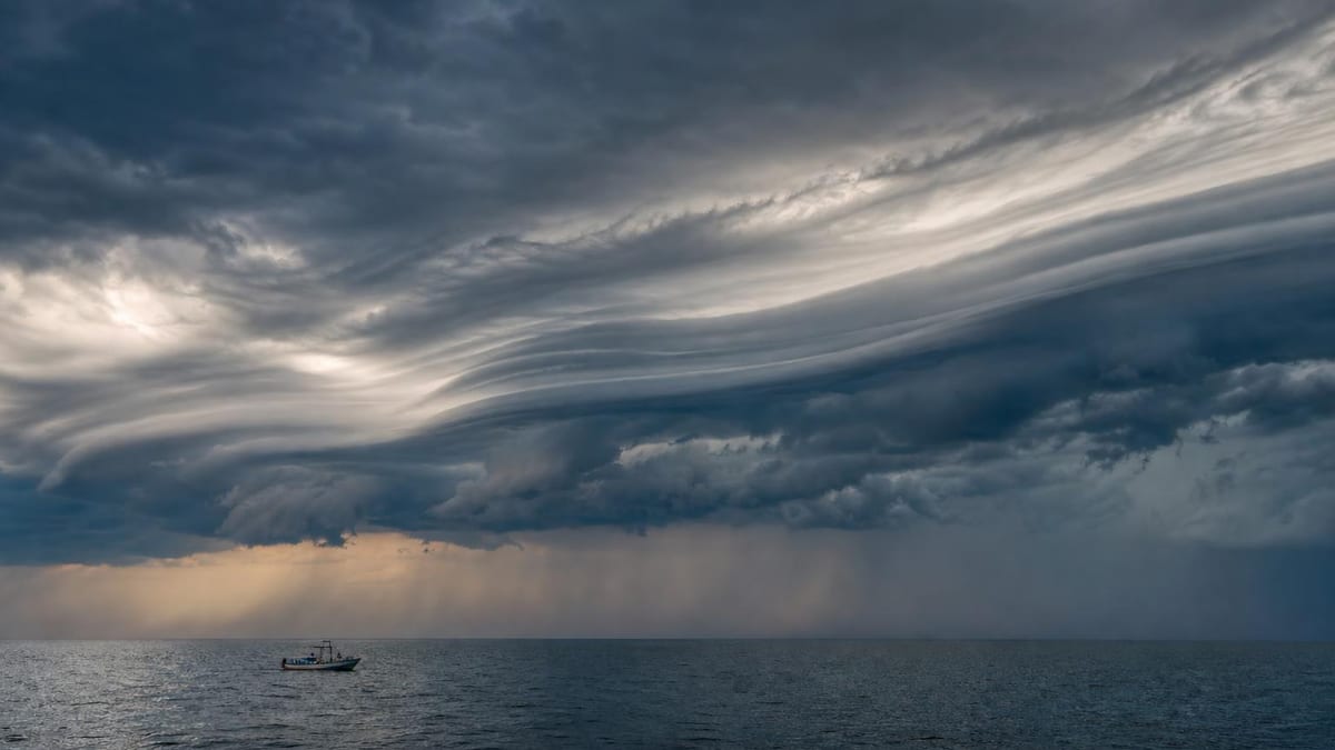 A small fishing boat drifts on a calm dark sea beneath a dramatic sky filled with layered gray storm clouds and distant rain