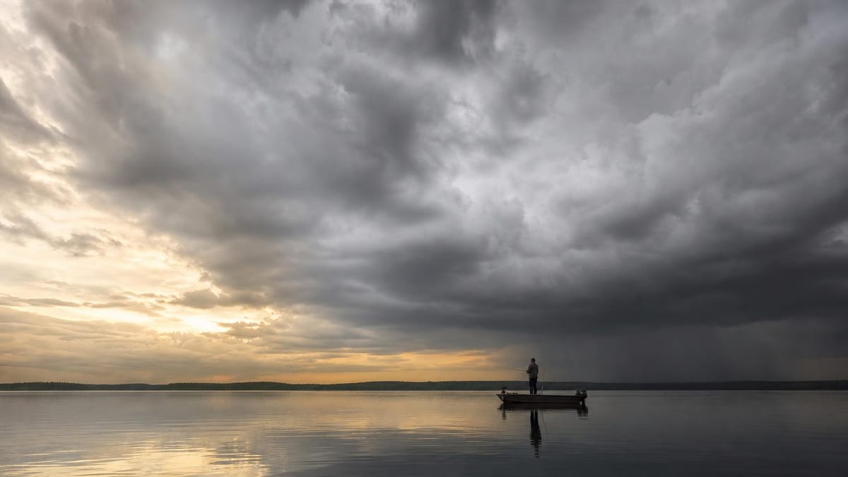 A lone angler stands in a small boat fishing on a calm lake at dusk beneath dramatic gray storm clouds, with warm sunset light glowing on the horizon and reflecting on the still water