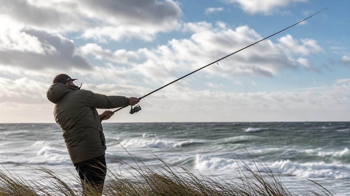 A man in an olive parka and cap casts a fishing rod with a spinning reel toward a rough, wave-filled sea while standing among tall coastal dune grass under a partly cloudy sky