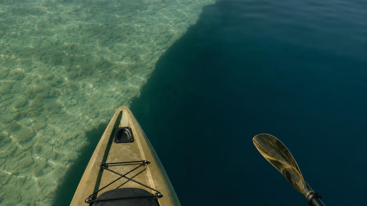 The bow of an olive-green kayak and a paddle blade extend over clear water that transitions from a shallow rocky bottom to deep dark blue