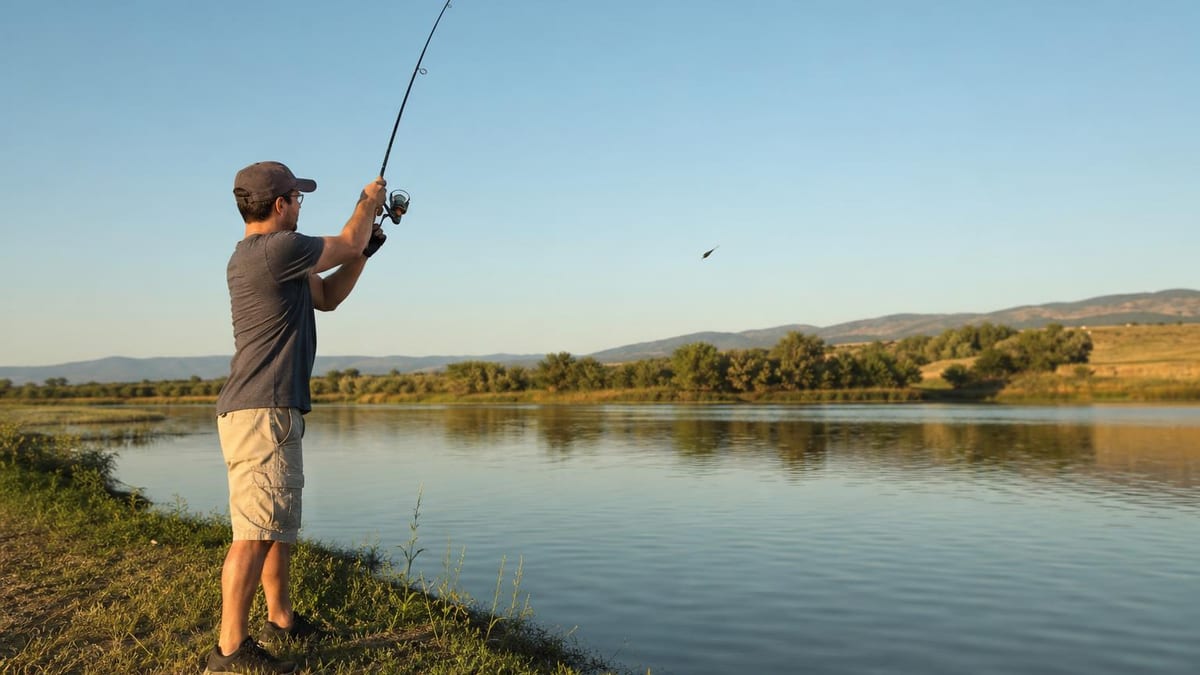 An angler on a grassy lake shore in late afternoon light at the release point of an overhead cast, spinning rod forward at eye level and the lure in flight over open water