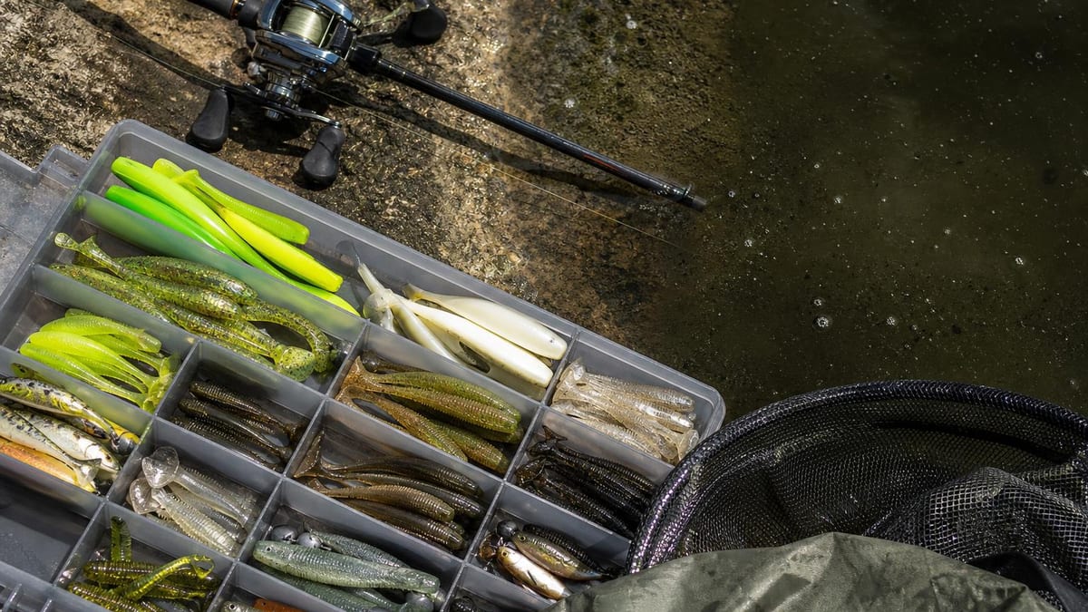 An open tackle box filled with various green, chartreuse, white, and natural-colored soft plastic fishing lures sits on a rocky shoreline beside a baitcasting reel and rod, a dark landing net, and calm water