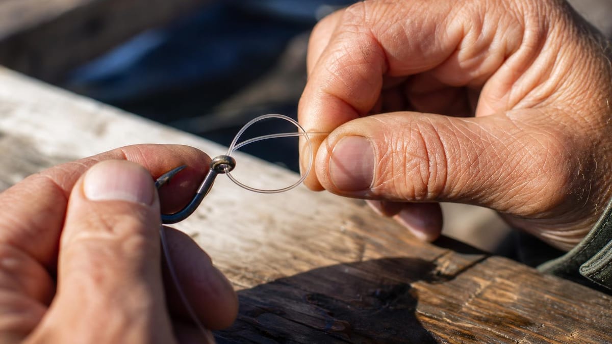 Close-up of a person's weathered hands threading a clear monofilament fishing line through the eye of a metal hook over a sunlit wooden surface